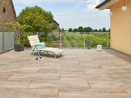 Ein Liegestuhl auf einer Terrasse mit Bodenplatten in Holzoptik, im Hintergrund ein Weinberg