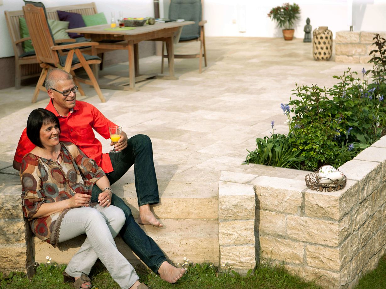 Besonders barfuß ein Genuss: Travertin Paar sitzt auf einer Terrasse mit Natursteinplatten aus Travertin Medium SELECT und Natursteinmauer, im Hintergrund Gartenmöbel aus Holz