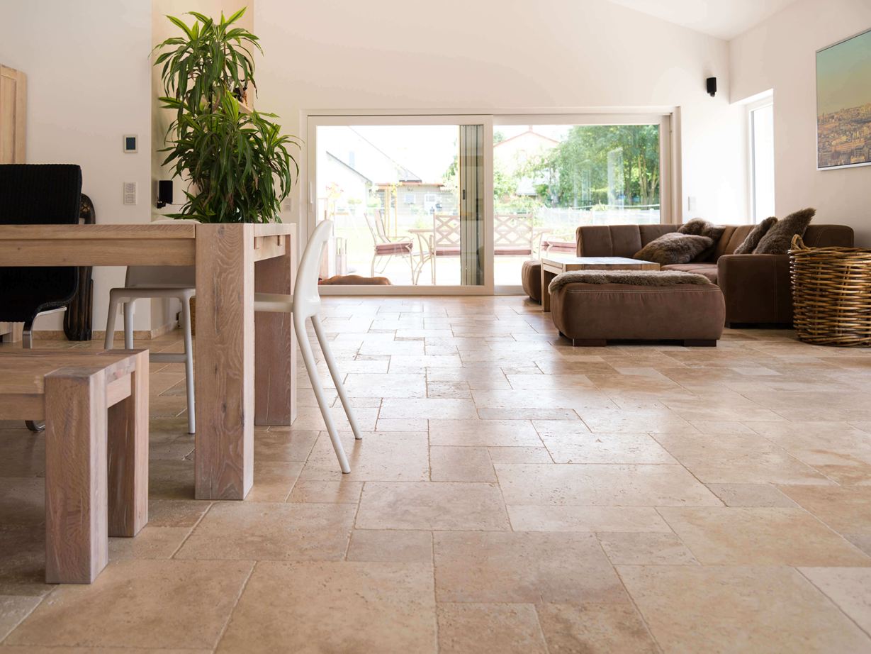 Die hellen Farbtöne wirken leicht und elegant. Wohnzimmer mit brauner Couch, weißen Wänden und Travertinfliesen Medium in Braun-Beige als Bodenbelag