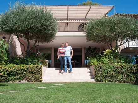 Paar steht vor einem Hotel mit Travertin als Bodenbelag, im Vordergrund eine Rasenfläche und links und rechts Bäume