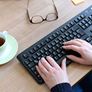Hände auf Keyboard, daneben eine Brille und eine Tasse Kaffee