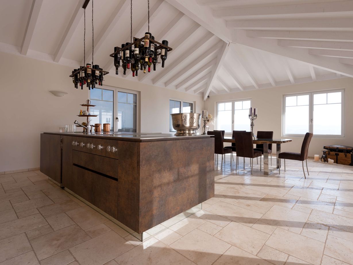 Kücheninsel mit Weinflaschenhalterung an der Decke, im Hintergrund Esstisch und Stühle auf verlegtem Travertin Light mit getrommelter, ungespachtelter Oberfläche