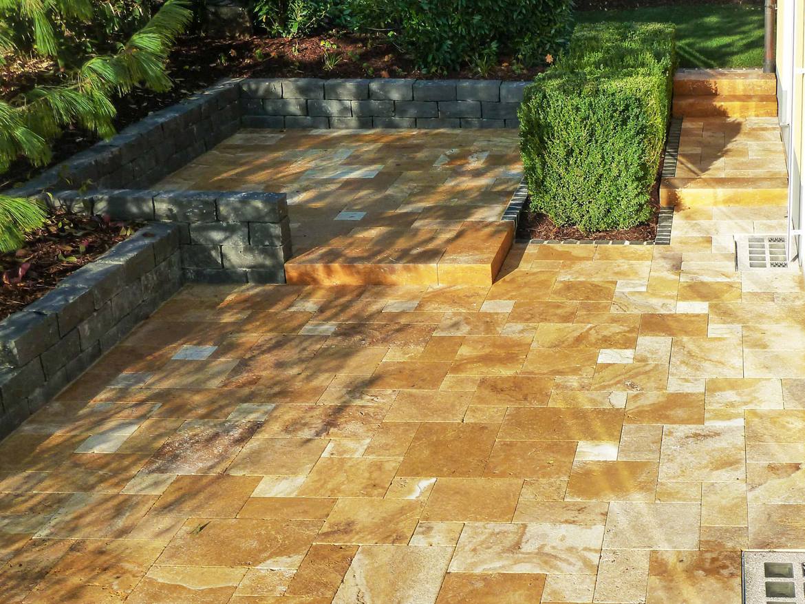 Terrassenfläche mit Travertinplatten Gold, niedriger grauer Mauer und grüner Hecke im Hintergrund