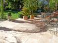 Terrasse mit Travertinplatte Medium Select, im Hintergrund Rasenfläche und mehrere Blumentöpfe, vorne rechts Gartenstühle mit bunten Kissen