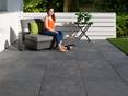 Verlegehinweise : Wir empfehlen, diese Feinsteinzeugplatten im Splittbett zu verlegen. Frau sitzt auf modernem Sessel auf Terrasse mit Platten in Schieferoptik