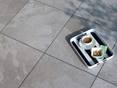 Diese Platten sind rutschfest und daher perfekt für die Terrasse. Tablett mit Kaffee auf Feinsteinzeugplatten in hellgrauer Schieferoptik Alpine Grey von oben fotografiert